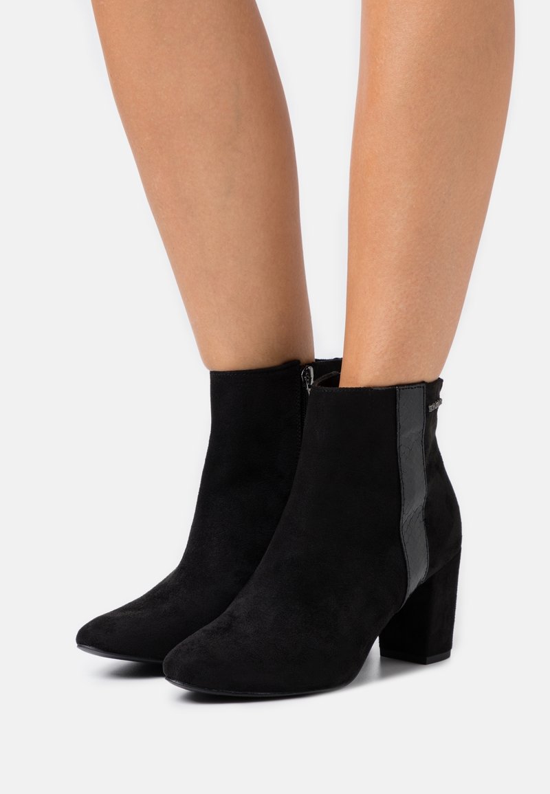 Bottines en daim noir à talons carrés, fermeture éclair latérale et détail de bande noire brillante sur les côtés extérieurs, portées sur jambes nues.