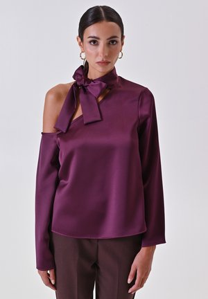 Blusa in satin viola con design a spalla scoperta, caratterizzata da un dettaglio del collo annodato e maniche lunghe. Texture liscia con una leggera lucentezza.