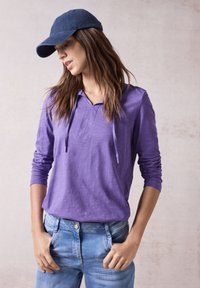 Felpa viola a maniche lunghe con colletto a camicia e lacci, abbinata a jeans in denim azzurri e un cappellino da baseball blu scuro.