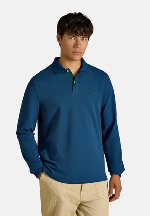 Giovane uomo che indossa una polo blu a maniche lunghe e pantaloni beige, in piedi con una mano in tasca davanti a uno sfondo semplice.