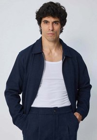 Veste zippée bleu marine avec un col, fabriquée en tissu texturé, portée sur un débardeur blanc. Comprend des poches avant et une coupe ajustée.