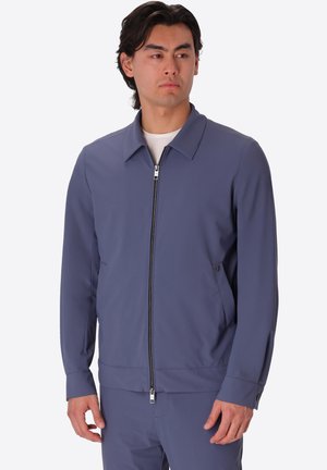 Uomo che indossa una giacca blu con zip e pantaloni abbinati, in piedi e che guarda di lato su uno sfondo semplice.
