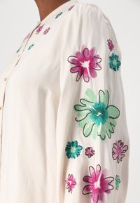 Krembluse med fargerik blomsterbroderi i rosa, grønn og blå, med avslappet passform og knappefeste foran.