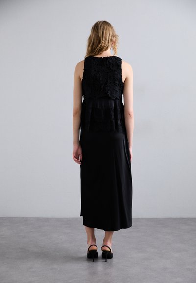 Mustmust kanna, millel on kihilised pitsdetailid rindkereosas ja sile satiinsärk. Mudel kannab musti kõrge kontsaga sandaale.