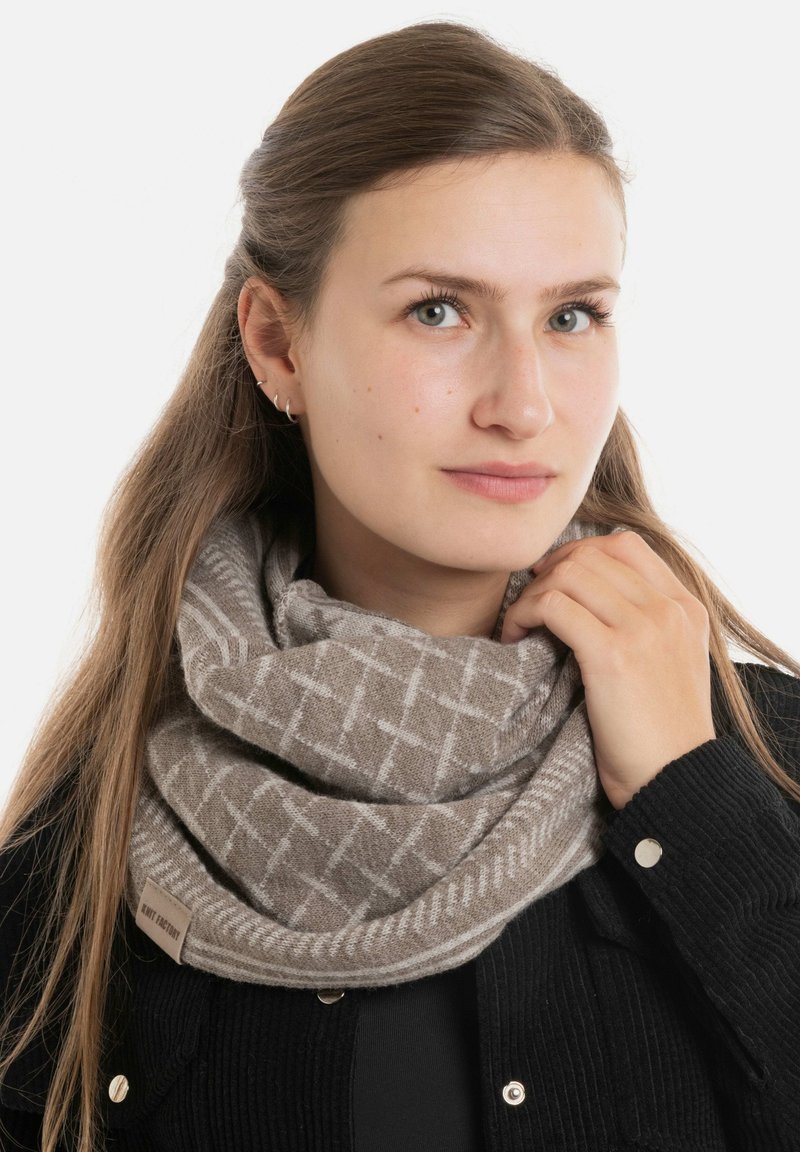 Gebreide bruine cowl-sjaal met een geometrisch patroon, zachte textuur en een lichtgrijze accent. Label zichtbaar aan de zijkant.