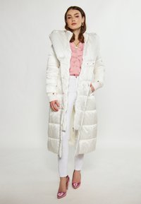 Hvid quiltet pufferjakke med oversize krave, parret med en pink bluse og hvide bukser. Har knaplukninger og ribbede manchetter, båret med skinnende pink heels.