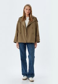 Chaqueta caqui de doble botonadura con amplios solapas, combinada con jeans de mezclilla azules y zapatillas blancas. La tela tiene una textura suave.
