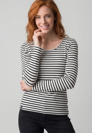 Red-haired woman with light skin smiling, wearing a black and white striped long-sleeve shirt and black pants, standing against a plain gray background.