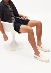 Białe gumowe chodaki z wycięciami po bokach, noszone z czarnymi szortami. Osoba siedzi w nowoczesnym białym fotelu na jasnym tle.
