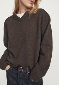 Hnědý pletený svetr s výstřihem do V a volným střihem, kombinovaný s tmavými džínami a hnědým koženým pásem. Měkká struktura s žebrovanými manžetami.