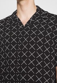 Schwarzes Kurzarmshirt mit einem weißen kreuzenden Rautenmuster, mit einem abgesenkten Kragen und einer Frontknopfleiste.