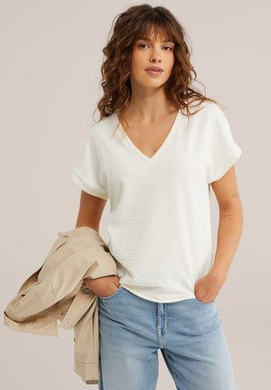 Tânără cu păr castaniu ondulat purtând un top alb texturat cu decolteu în V și blugi albastru deschis, ținând o jachetă bej într-o mână.