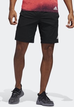 Mężczyzna ubrany w czarne spodenki Adidas, czarno-szare buty sportowe oraz czerwoną koszulkę Adidas, stojący z lekko rozstawionymi nogami.