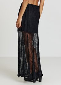 Jupe en dentelle noire avec une fente haute, une texture semi-transparente et une taille ajustée. L'ourlet arrive jusqu'au sol, mettant en valeur la visibilité des jambes.