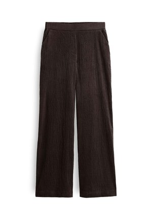 Nahaufnahme einer Person, die dunkelbraune, hoch taillierte, gerippte Hose und ein weißes Hemd trägt, mit den Händen auf den Hüften.