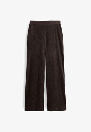 Nahaufnahme einer Person, die dunkelbraune, hoch taillierte, gerippte Hose und ein weißes Hemd trägt, mit den Händen auf den Hüften.