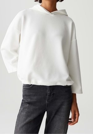 Personne portant un haut à capuche blanc texturé à manches longues avec un ourlet élastique et un jean gris foncé, visible du cou jusqu'à mi-cuisse.