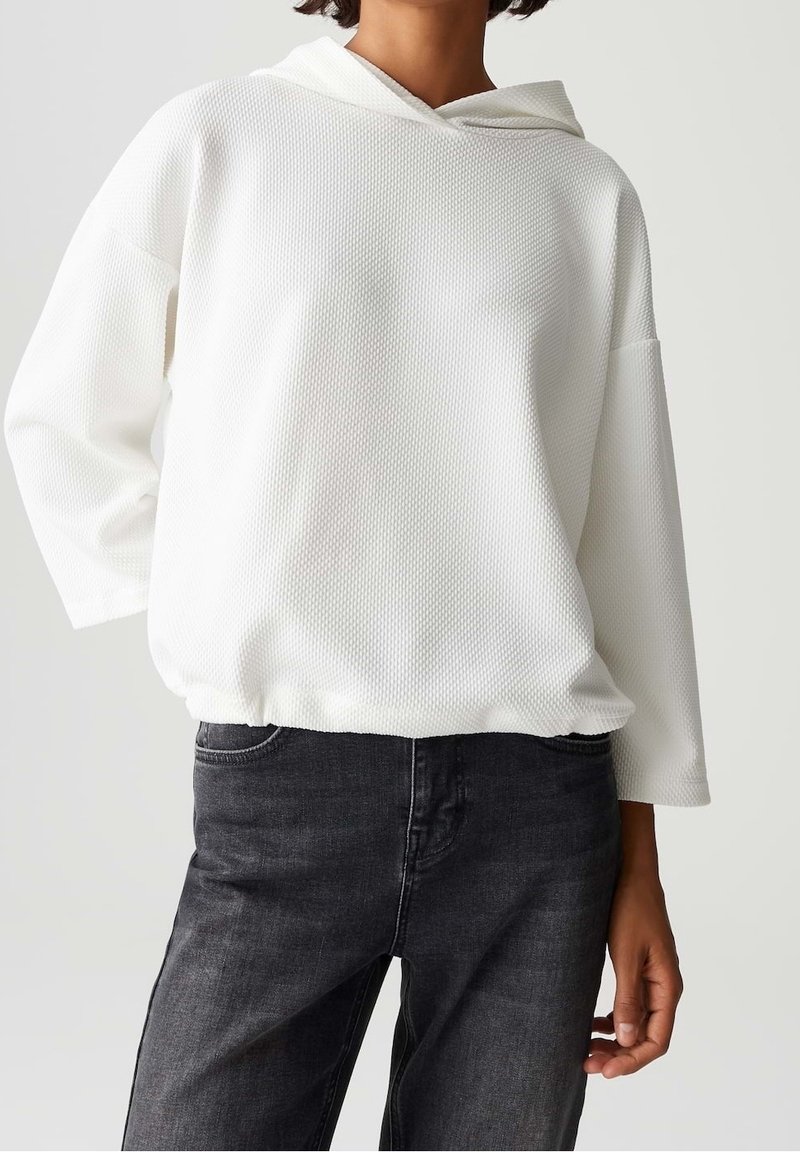 Personne portant un haut à capuche blanc texturé à manches longues avec un ourlet élastique et un jean gris foncé, visible du cou jusqu'à mi-cuisse.
