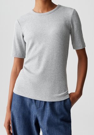 Personne portant un haut à manches courtes côtelé gris clair et un pantalon bleu texturé, une main dans la poche, sur un fond uni.