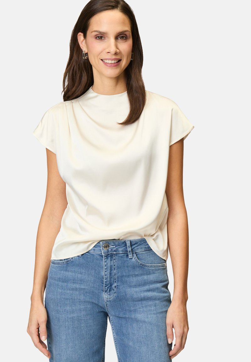 Helles beige Kurzarmoberteil mit hohem Halsausschnitt und glatter Textur, kombiniert mit klassischen blauen Denim-Jeans mit Knopfverschluss.