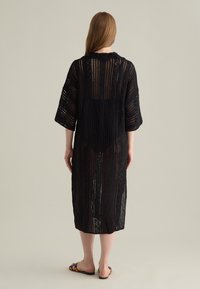 Schwarzes gestricktes Überkleid mit dreiviertel Ärmeln, offenem Gewebe-Muster, knielang und runder Ausschnitt. Getragen mit bunten Sandalen.