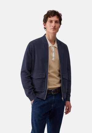 Uomo che indossa una giacca blu navy con zip sopra una polo beige e jeans blu con cintura marrone, in piedi davanti a uno sfondo semplice.