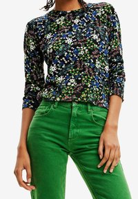 Kobieta w czarnej bluzce w kwiaty z długim rękawem, połączonej z jasnymi zielonymi spodniami z wysokim stanem, widoczna od ramion do bioder.