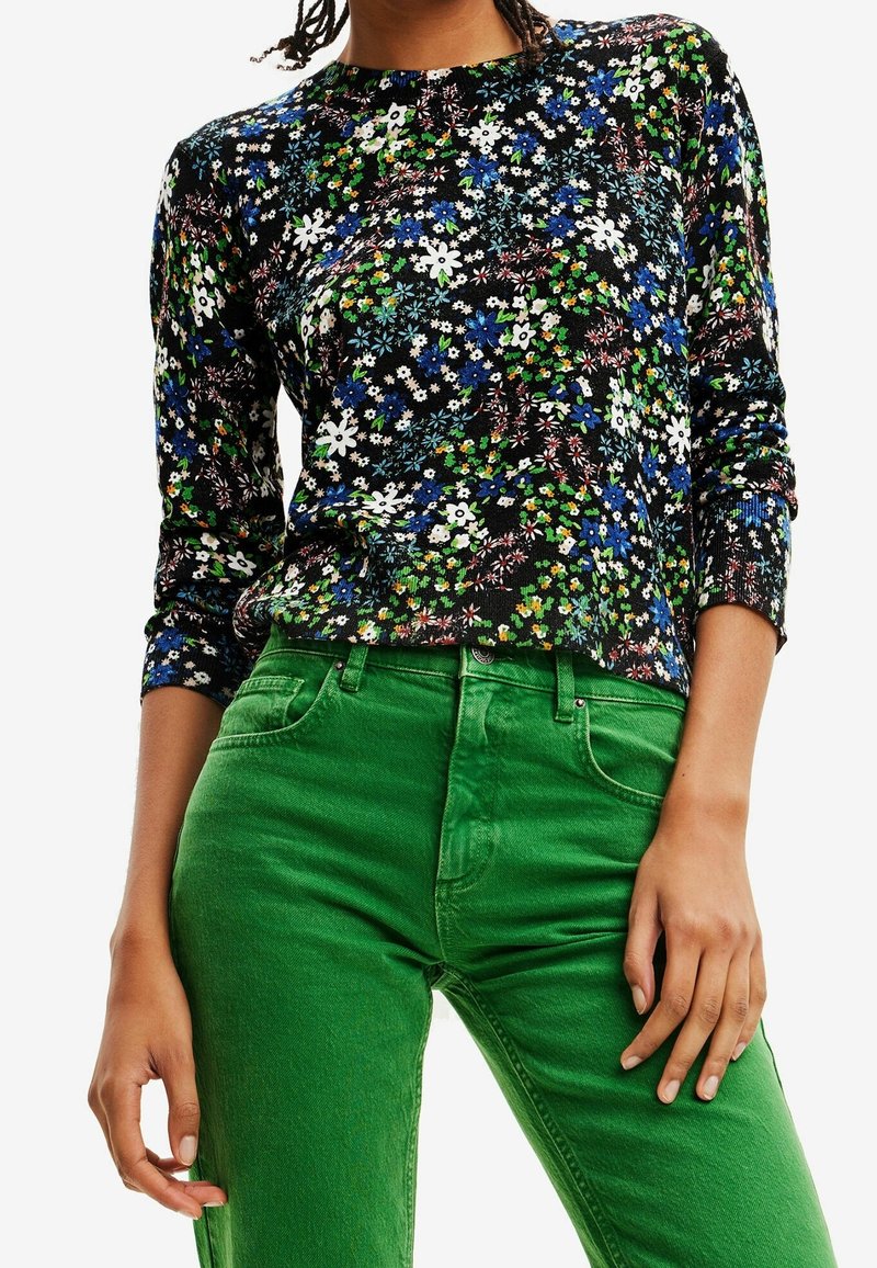 Kobieta w czarnej bluzce w kwiaty z długim rękawem, połączonej z jasnymi zielonymi spodniami z wysokim stanem, widoczna od ramion do bioder.
