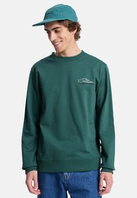 Sudadera verde de manga larga, con puños acanalados y cuello redondo. Presenta un logo blanco en el lado izquierdo del pecho. Se lleva con pantalones de mezclilla azul.