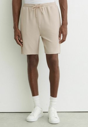 Uomo con pantaloncini beige a trama con coulisse, calzini bianchi al polpaccio e scarpe da ginnastica bianche, in piedi contro una parete bianca semplice.