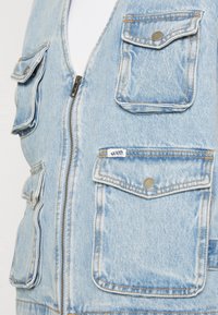 Ljusblå denimväst med flera knäppta fickor och en framsliten dragkedja, med en liten vit Guess Originals-tagg.