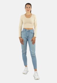 Beige langärmliger Cropped-Top, kombiniert mit hochgeschnittenen hellblauen Jeans und weißen Sneakern. Das Model steht mit den Händen in den Hüften.
