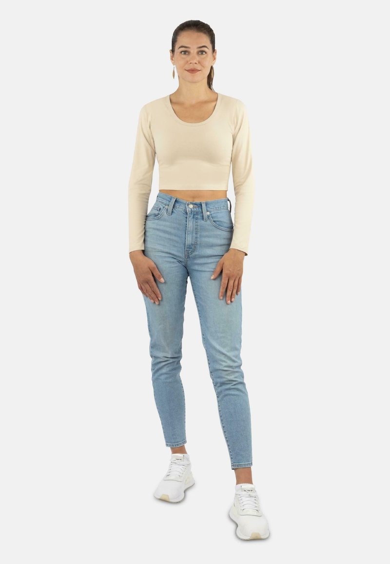 Beige langärmliger Cropped-Top, kombiniert mit hochgeschnittenen hellblauen Jeans und weißen Sneakern. Das Model steht mit den Händen in den Hüften.
