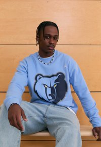 Világoskék pulóver sötétkék és szürke medve grafikával. Kerek nyakkivágással és bordázott mandzsettákkal rendelkezik. Világoskék farmerrel viselve.