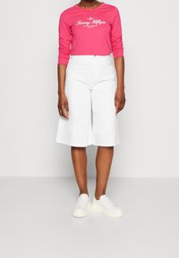 Camicia a maniche lunghe rosa con logo bianco "Tommy Hilfiger", abbinata a pantaloni bianchi in stile culotte e sneakers bianche con platform.
