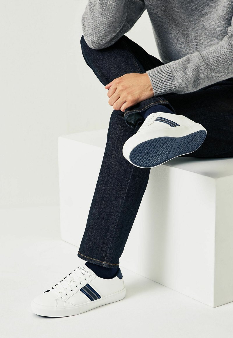 Weiße Sneakers mit maritimen Streifen, Schnürdesign, Gummisohle und runder Zehenpartie. Getragen mit dunklen Denim-Jeans und einem hellgrauen Pullover.