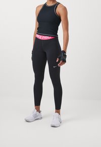 Persona con una camiseta sin mangas negra, leggings Nike Pro negros con cinturilla rosa, guantes sin dedos y zapatillas Nike blancas de pie.