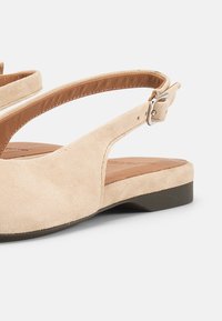 Μπεζ σανδάλι φλατ με λουράκι λουστρίνι, ρυθμιζόμενο ασημί αγκράφα και καφέ μαλακό εσωτερικό σε ανοιχτό φόντο.