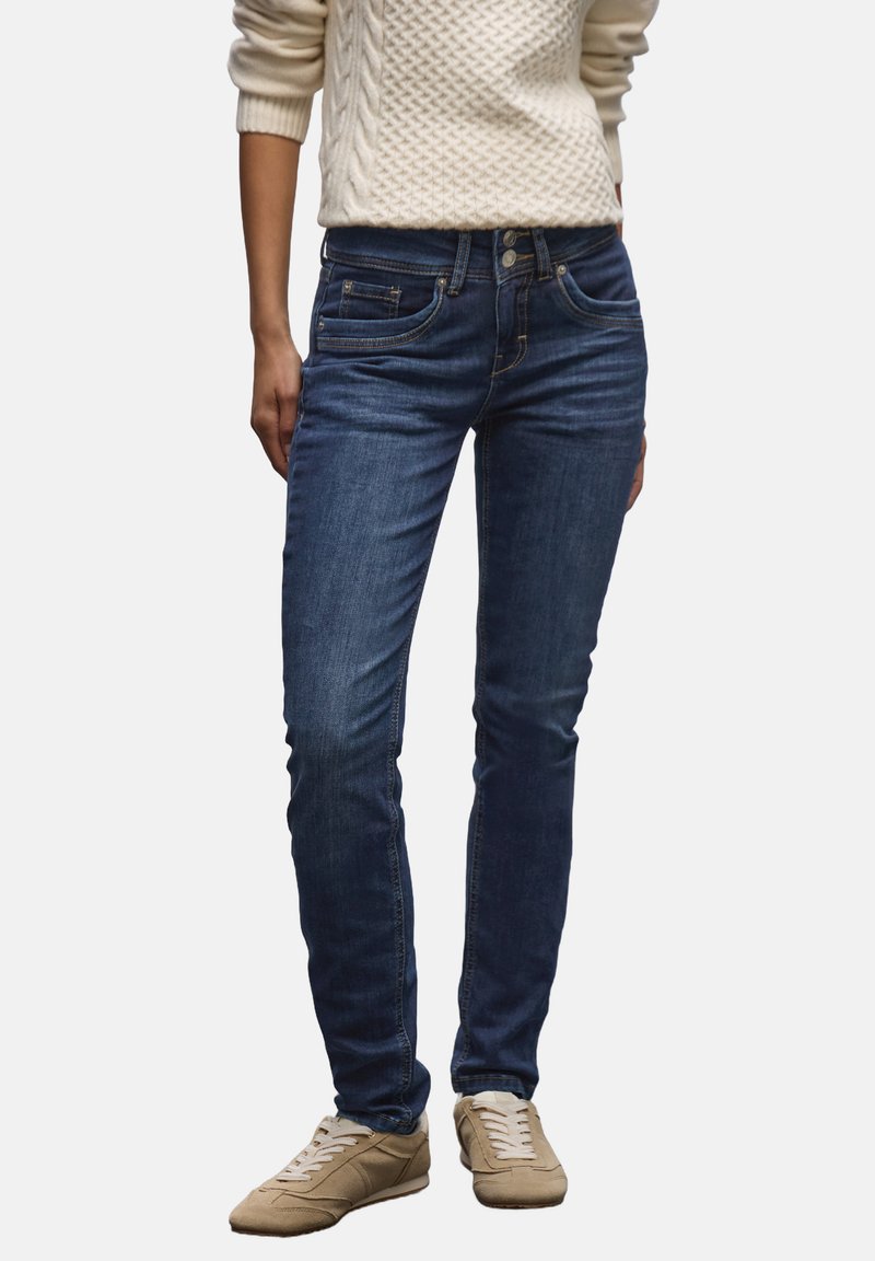 Vaqueros de denim azul oscuro con corte ajustado, diseño de cinco bolsillos y detalles de costura visibles, combinados con un suéter de punto trenzado en crema y zapatos beige.