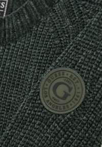 Maglione verde scuro in tessuto a rete testurizzata; presenta una toppa rotonda con logo in rilievo in toni tenui sul lato sinistro.