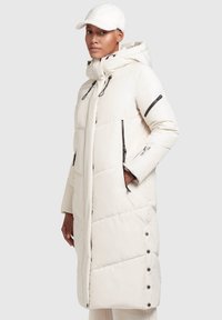 Femme portant un long manteau blanc rembourré avec des zippers et des détails à pression noirs, associé à un bonnet texturé blanc, sur un fond uni.
