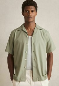 Camisa de manga corta, con botones, en tejido verde claro, que presenta un cuello tipo campamento y una textura suave, combinada con una camiseta blanca por debajo.