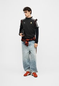 Νεαρός άνδρας που φοράει μαύρο πουκάμισο Puma Ferrari πάνω α πό μακρυμάνικη μπλούζα, ανοιχτόχρωμο μπλε φαρδιά τζιν, κόκκινα αθλητικά παπούτσια, γυαλιά και κρατάει μια μικρή μαύρη τσάντα.