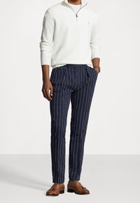 Vit dragkedja tröja och marinblå randiga byxor. Byxorna har plisserad front och bälteshällor, kompletterat av bruna loafers.