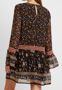 Vestido negro de flores con mangas acampanadas largas, ribete geométrico rojo y beige en la cintura, puños y dobladillo, llevado por una persona vista desde atrás.