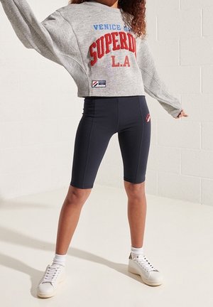 Person iført en grå cropped sweatshirt med "SUPERDRY" i rødt, navy cykelshorts, hvide sokker og hvide sneakers, stående mod en hvid væg.