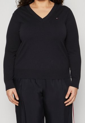 Kvinde iført en sort V-hals langærmet sweater og matchende sorte bukser med et lille logo på brystet.