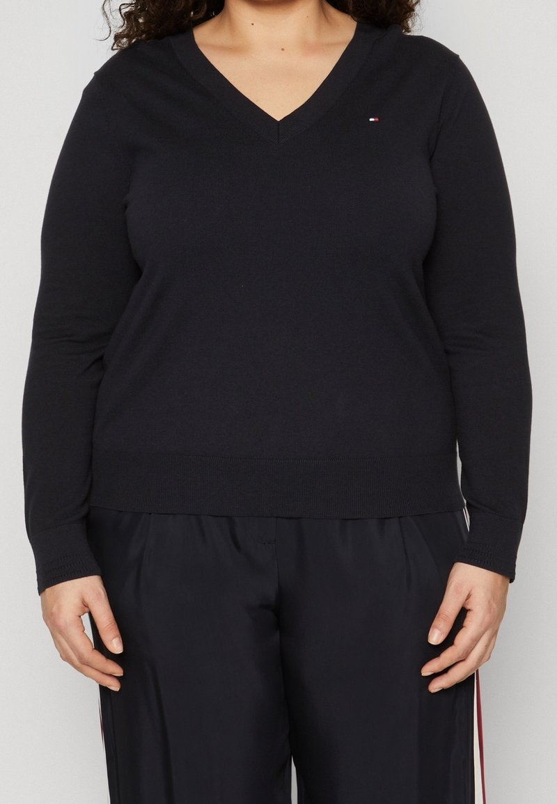 Femme portant un pull noir à col en V manches longues et un pantalon noir assorti avec un petit logo sur la poitrine.