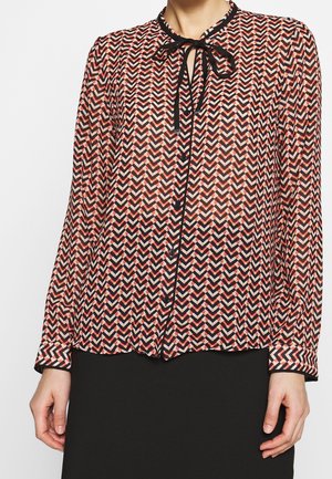 Bluzka z długim rękawem w czerwono-czarno-biały wzór w jodełkę, z czarnymi guzikami z przodu i czarną kokardą przy dekolcie, noszona z czarną spódnicą.