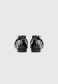 Pair of shiny black leather flat shoes with ankle straps and buckles, viewed from the back on a plain white background.